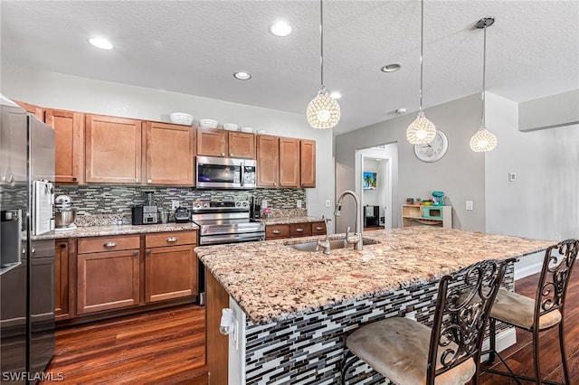 kitchen with sink, a center island with sink, pendant lighting, stainless steel appliances, and light stone countertops