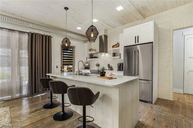 kitchen featuring stainless steel refrigerator, sink, hanging light fixtures, wall chimney range hood, and an island with sink