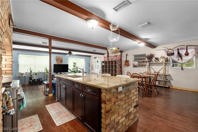 kitchen with sink, dark wood-type flooring, beam ceiling, light stone counters, and an island with sink