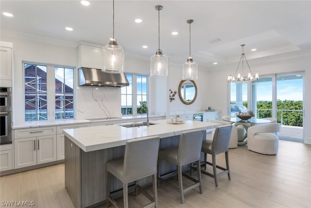 kitchen featuring hanging light fixtures, light stone countertops, range hood, a kitchen island with sink, and a sink