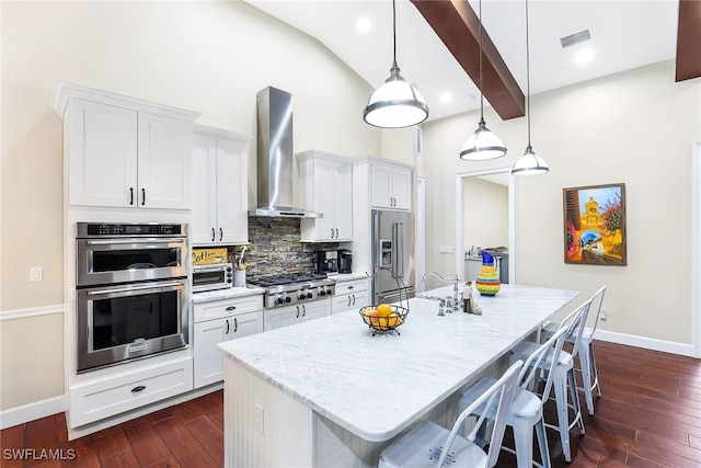 kitchen featuring white cabinets, wall chimney exhaust hood, decorative light fixtures, stainless steel appliances, and a kitchen island with sink