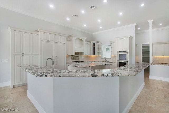 kitchen with light stone counters, stainless steel double oven, a large island with sink, and tasteful backsplash