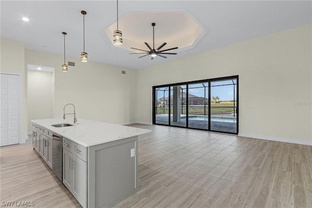 kitchen featuring pendant lighting, sink, light stone counters, a center island with sink, and a raised ceiling