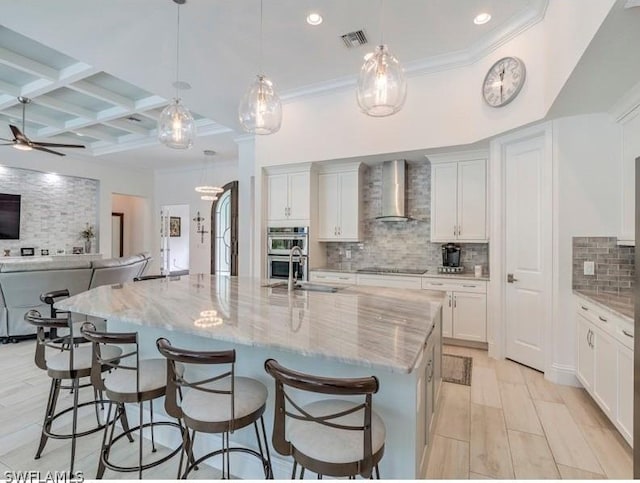 kitchen with coffered ceiling, ceiling fan, wall chimney range hood, a large island with sink, and beamed ceiling