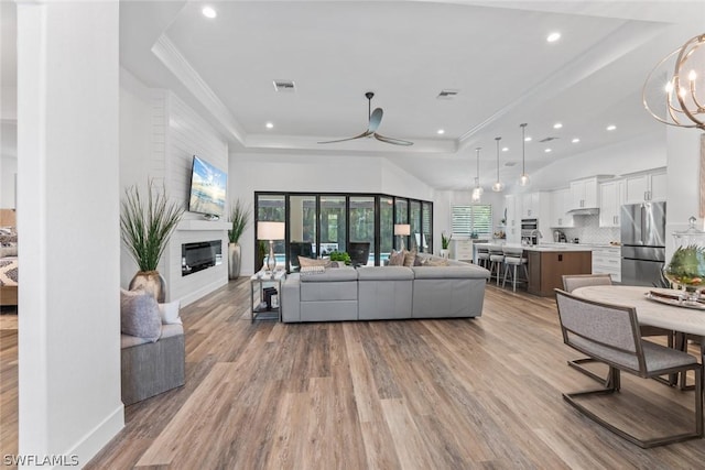 living area with visible vents, a raised ceiling, a glass covered fireplace, crown molding, and light wood-type flooring