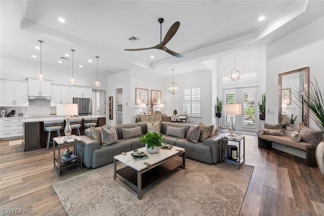 living room with ceiling fan with notable chandelier, wood-type flooring, ornamental molding, and a raised ceiling