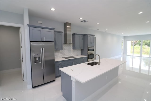 kitchen featuring light stone countertops, sink, wall chimney range hood, an island with sink, and appliances with stainless steel finishes