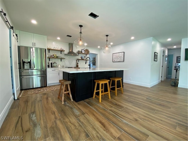 kitchen featuring stainless steel refrigerator with ice dispenser, wall chimney range hood, a barn door, white cabinetry, and an island with sink