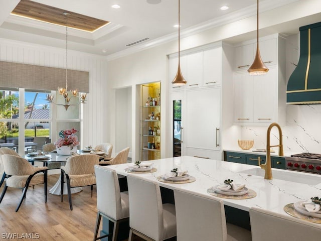 kitchen featuring white cabinetry, a raised ceiling, decorative light fixtures, wall chimney range hood, and light stone counters