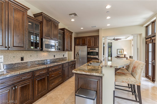 kitchen with a kitchen bar, stainless steel appliances, ornate columns, an island with sink, and dark brown cabinets