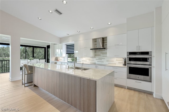 kitchen featuring white cabinets, vaulted ceiling, wall chimney exhaust hood, stainless steel double oven, and a kitchen island with sink