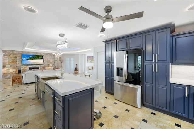 kitchen featuring appliances with stainless steel finishes, a tray ceiling, a kitchen island with sink, blue cabinets, and ceiling fan with notable chandelier