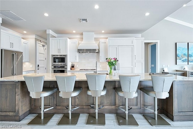 kitchen with stainless steel appliances, white cabinets, custom range hood, a large island with sink, and a breakfast bar