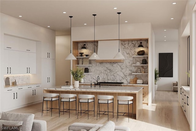 kitchen featuring light wood-type flooring, a center island with sink, premium range hood, white cabinets, and pendant lighting