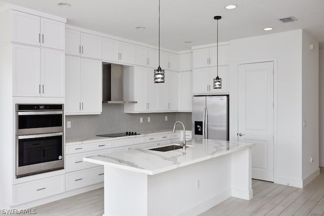 kitchen featuring white cabinetry, light stone countertops, wall chimney exhaust hood, a kitchen island with sink, and appliances with stainless steel finishes