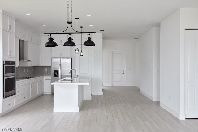 kitchen featuring a center island with sink, white cabinets, wall chimney range hood, hanging light fixtures, and light hardwood / wood-style flooring