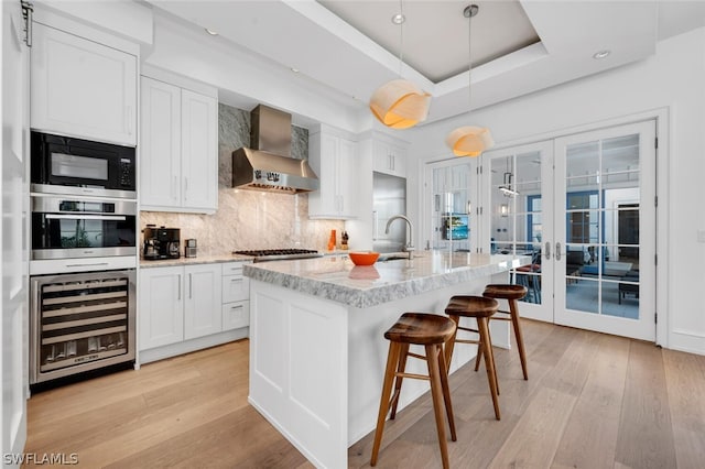 kitchen featuring wall chimney range hood, hanging light fixtures, stainless steel appliances, white cabinets, and beverage cooler