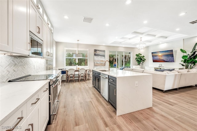 kitchen with coffered ceiling, white cabinetry, stainless steel appliances, decorative light fixtures, and a kitchen island with sink