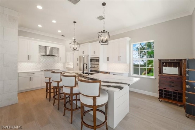 kitchen featuring stainless steel appliances, white cabinetry, wall chimney range hood, and an island with sink