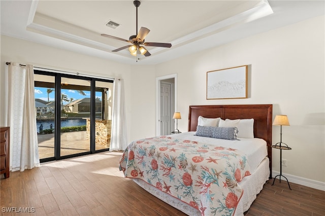 bedroom with hardwood / wood-style floors, access to exterior, ceiling fan, a tray ceiling, and a water view