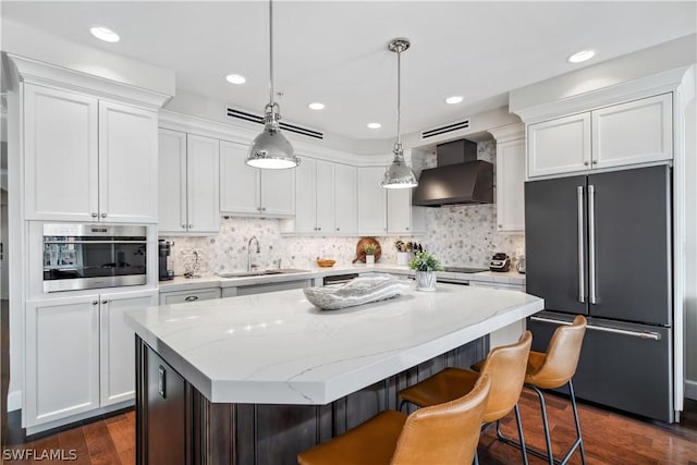 kitchen with wall chimney range hood, stainless steel appliances, a center island, white cabinets, and decorative light fixtures