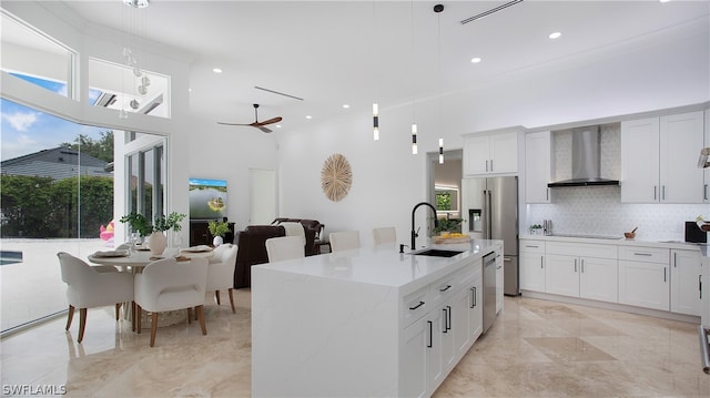 kitchen with hanging light fixtures, a center island with sink, wall chimney range hood, white cabinets, and appliances with stainless steel finishes