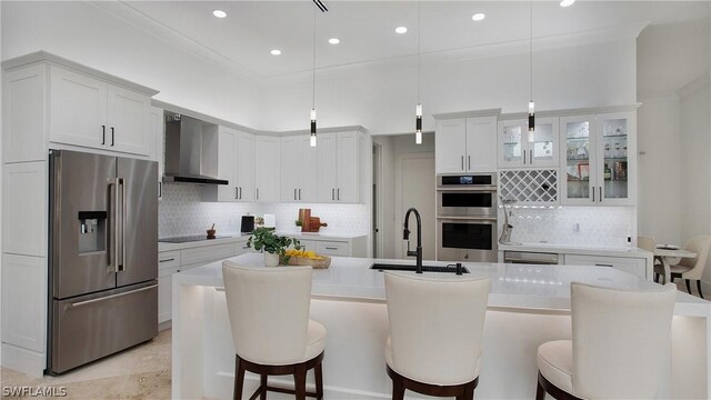 kitchen featuring a center island with sink, stainless steel appliances, pendant lighting, white cabinetry, and wall chimney range hood