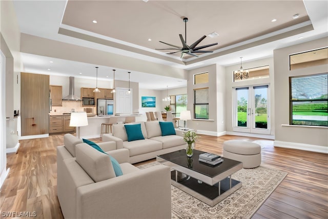 living room featuring french doors, a raised ceiling, light wood-type flooring, and ceiling fan with notable chandelier