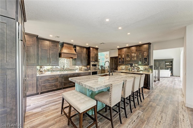 kitchen featuring custom range hood, a textured ceiling, light wood-type flooring, a center island with sink, and sink