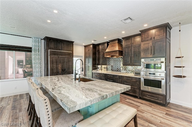 kitchen featuring an island with sink, custom exhaust hood, light hardwood / wood-style flooring, double oven, and sink