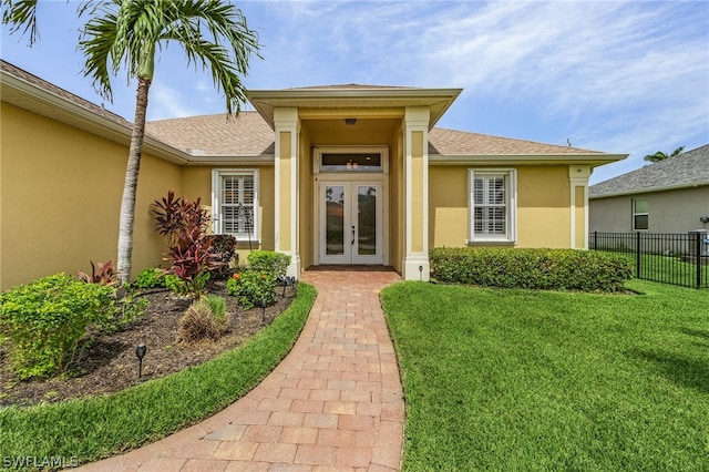 entrance to property with stucco siding, a lawn, fence, french doors, and a shingled roof