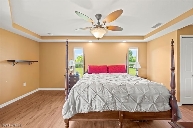 bedroom with ceiling fan, light hardwood / wood-style flooring, a tray ceiling, and multiple windows