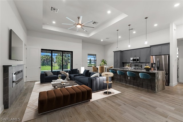 living room with a towering ceiling, light wood-type flooring, a raised ceiling, ceiling fan, and a tiled fireplace