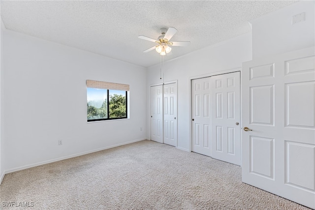 unfurnished bedroom with ceiling fan, a textured ceiling, light carpet, baseboards, and two closets