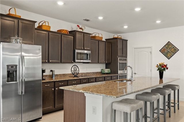 kitchen with dark brown cabinets, stainless steel appliances, an island with sink, sink, and light stone countertops