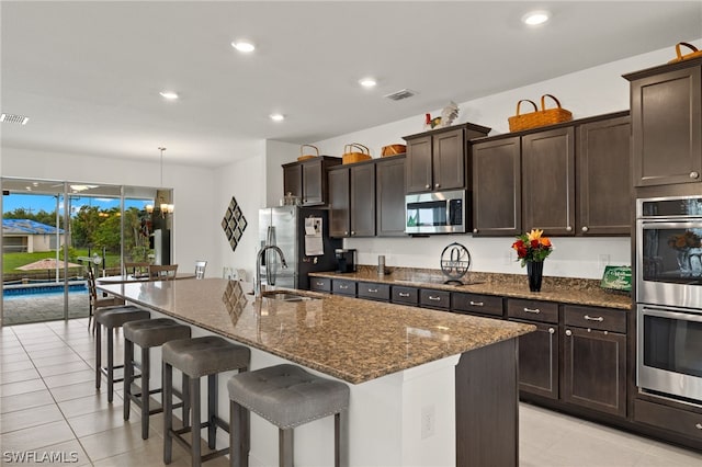 kitchen featuring stainless steel appliances, dark brown cabinets, pendant lighting, an island with sink, and light tile patterned flooring