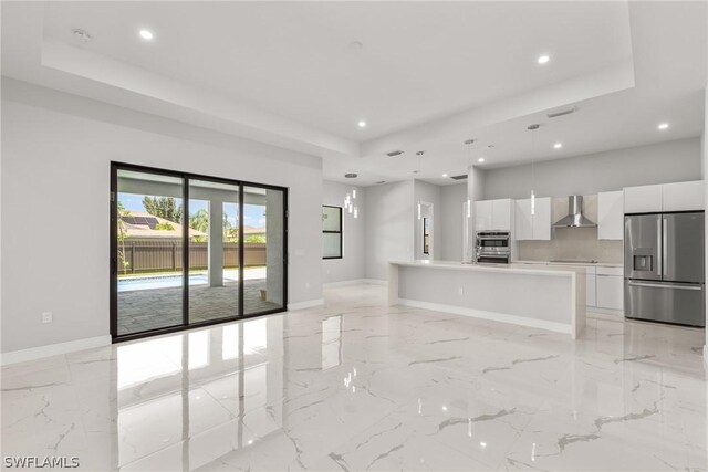 kitchen with wall chimney range hood, a center island with sink, a raised ceiling, stainless steel refrigerator with ice dispenser, and white cabinetry