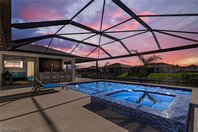 pool at dusk with ceiling fan, glass enclosure, exterior bar, a patio area, and an in ground hot tub