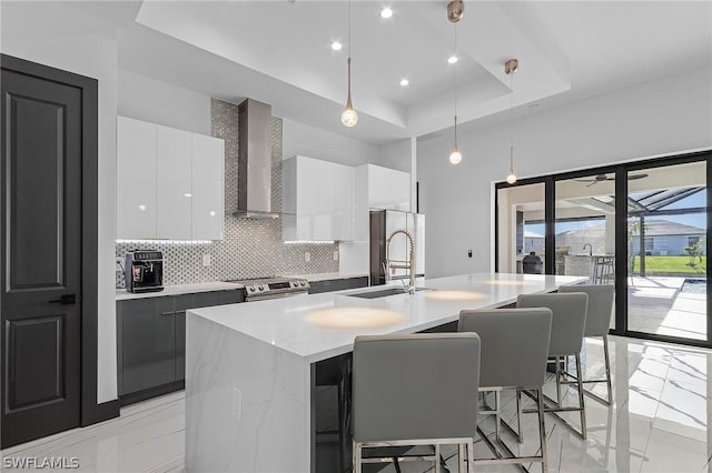 kitchen featuring pendant lighting, wall chimney range hood, white cabinetry, a tray ceiling, and an island with sink