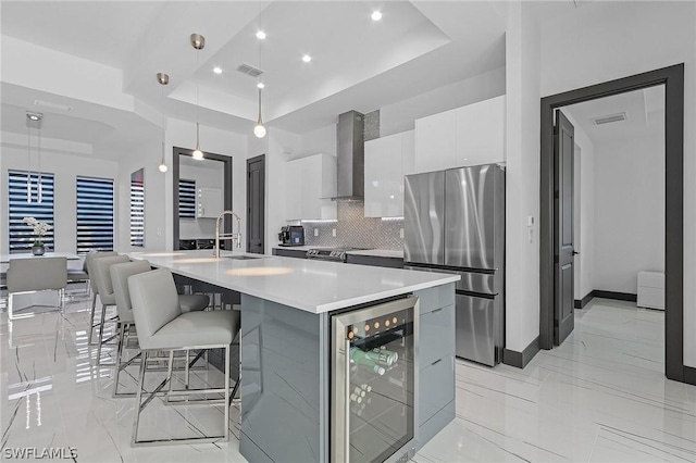 kitchen featuring wall chimney range hood, appliances with stainless steel finishes, a kitchen island with sink, a raised ceiling, and beverage cooler