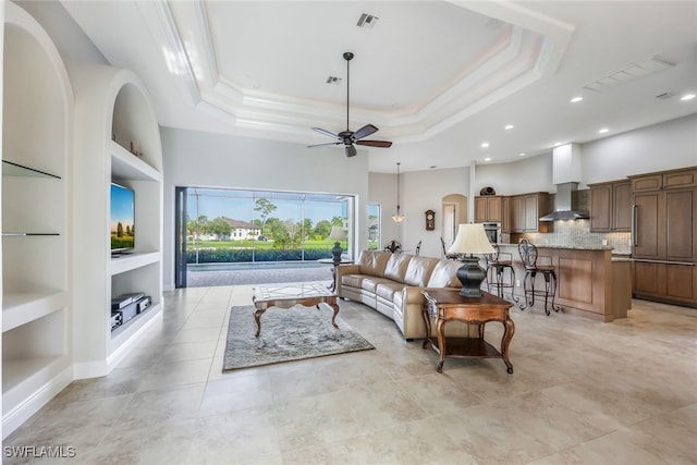 tiled living room with a raised ceiling, crown molding, built in features, and ceiling fan