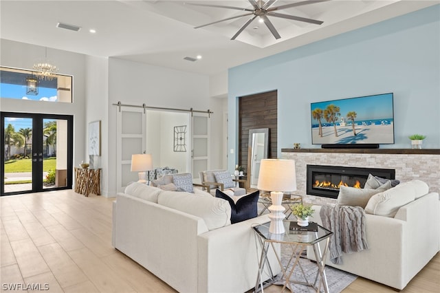 living room featuring french doors, a stone fireplace, a barn door, ceiling fan with notable chandelier, and light wood-type flooring