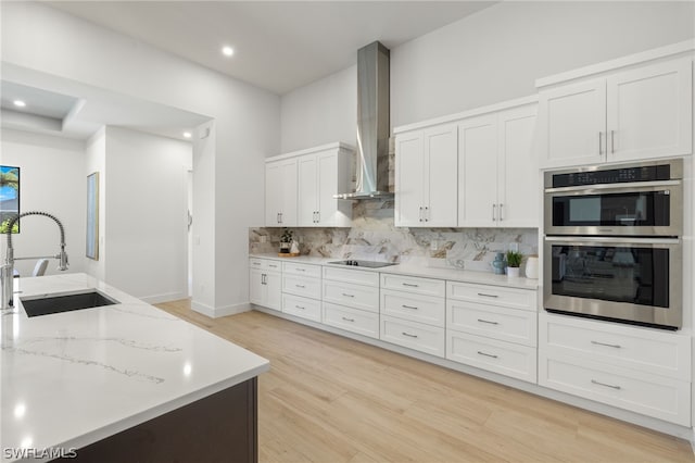 kitchen with wall chimney range hood, sink, double oven, tasteful backsplash, and white cabinetry