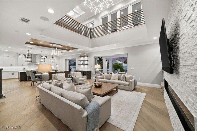 living room featuring a towering ceiling, a tray ceiling, light hardwood / wood-style flooring, an inviting chandelier, and a stone fireplace