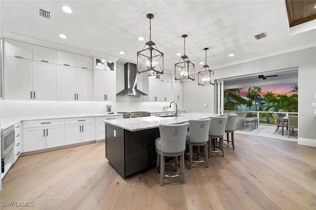 kitchen featuring decorative light fixtures, an island with sink, white cabinetry, and wall chimney range hood