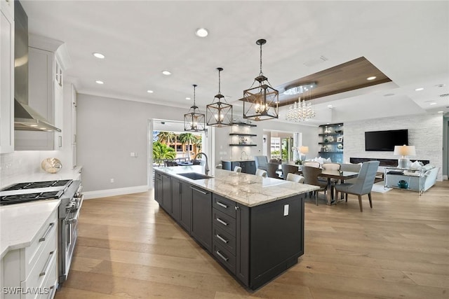 kitchen featuring wall chimney range hood, pendant lighting, high end stainless steel range, white cabinets, and an island with sink