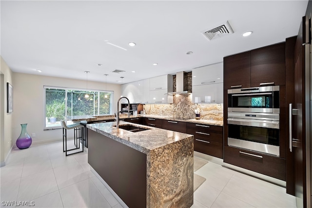 kitchen with backsplash, a kitchen island with sink, white cabinets, wall chimney range hood, and decorative light fixtures