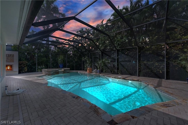 pool at dusk with an in ground hot tub, glass enclosure, exterior fireplace, and a patio area