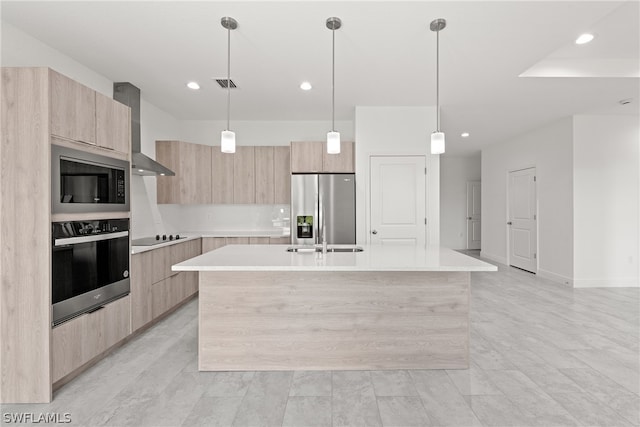 kitchen featuring wall chimney range hood, light brown cabinetry, hanging light fixtures, an island with sink, and stainless steel appliances