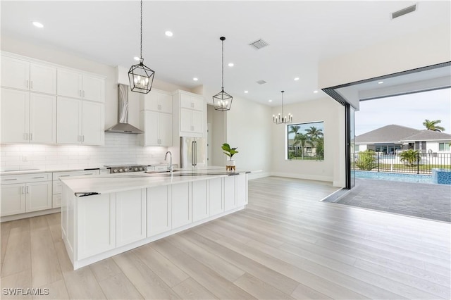 kitchen with white cabinets, wall chimney exhaust hood, pendant lighting, and an island with sink